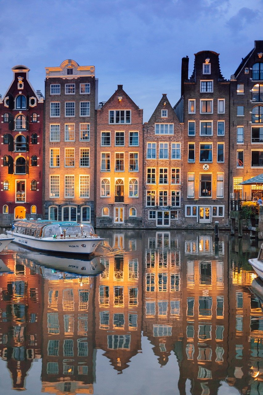 Historische grachtenpanden met verlichte ramen die reflecteren op het water in Amsterdam tijdens de schemering met een boot op de voorgrond