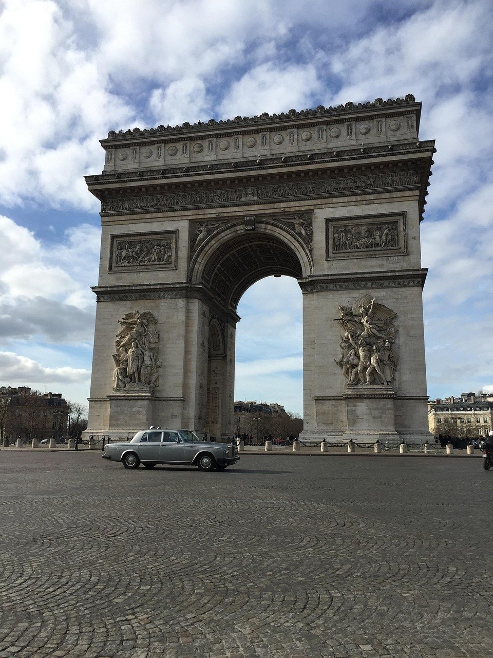 Arc de Triomphe in Parijs met een vintage zilveren auto geparkeerd op een geplaveid plein onder een deels bewolkte hemel