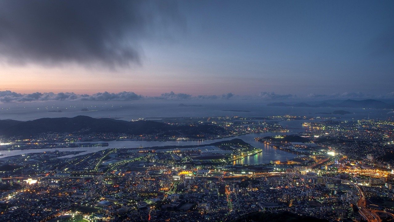 Luchtfoto van een kuststad bij schemering met verlichte gebouwen, waterwegen en verre bergen onder een bewolkte hemel