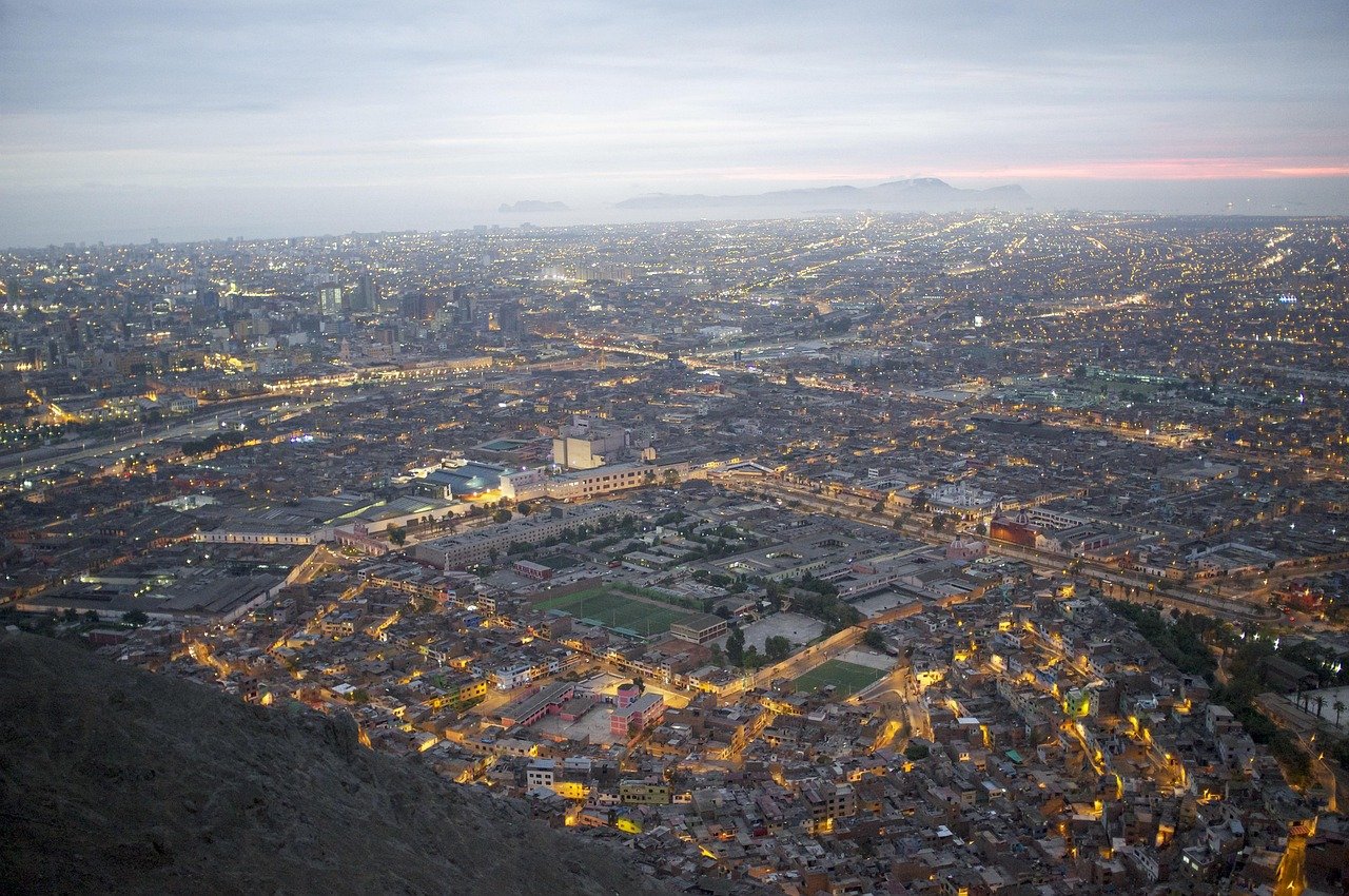 Luchtfoto van een uitgestrekte stadsgezicht bij schemering met verlichte straten en verre bergen onder een bewolkte hemel