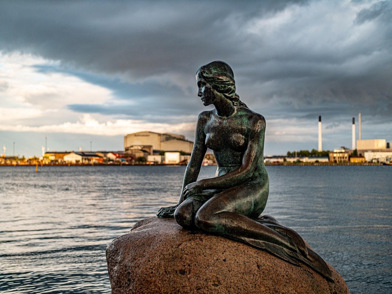 Het standbeeld van de Kleine Zeemeermin zittend op een rots bij het water in Kopenhagen met industriële gebouwen en bewolkte lucht op de achtergrond