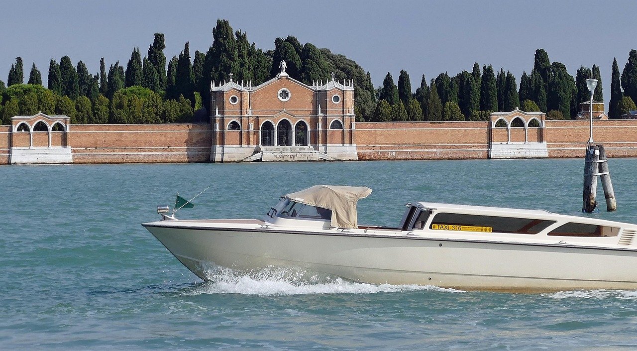 Witte watertaxi boot varend op turquoise water nabij historisch bakstenen gebouw en hoge cipressen
