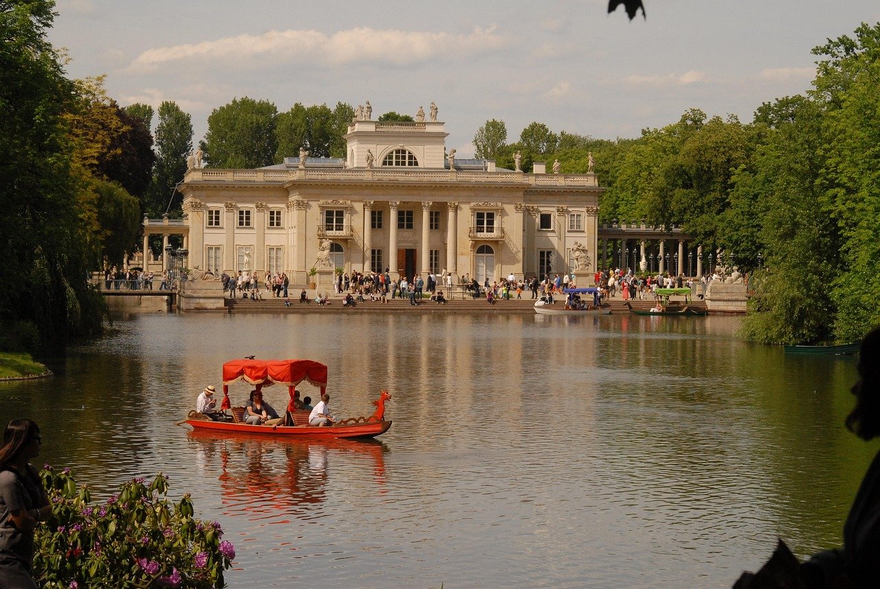 Historisch paleis met klassieke architectuur aan een meer met een rode boot en toeristen die genieten van een zonnige dag in een groen park
