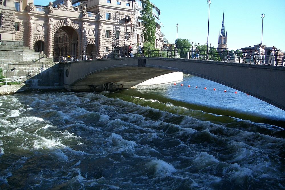 Stenen boogbrug over rivier met historische gebouwen Stenen boogbrug over woelig rivierwater met historische gebouwen en kerktoren op de achtergrond onder een heldere hemel