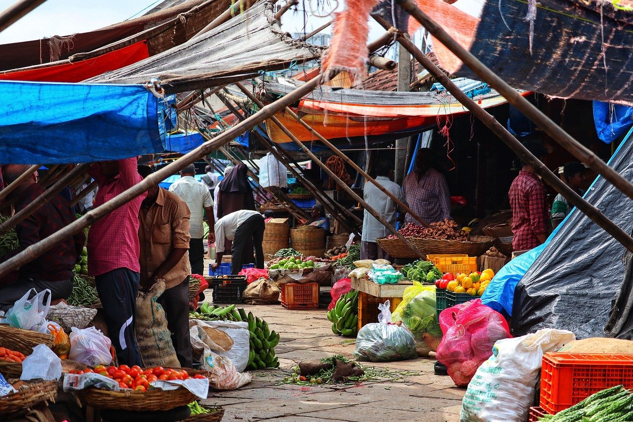 Drukke buitenmarkt met verse groenten, fruit en mensen die winkelen onder kleurrijke zeilen in een levendige straatomgeving