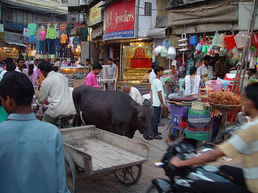 Drukke Indiase straatmarkt met buffel en winkels Drukke Indiase straatmarkt met mensen, een buffel, motorfiets en verschillende winkels, waaronder een juwelier en plasticwarenwinkel