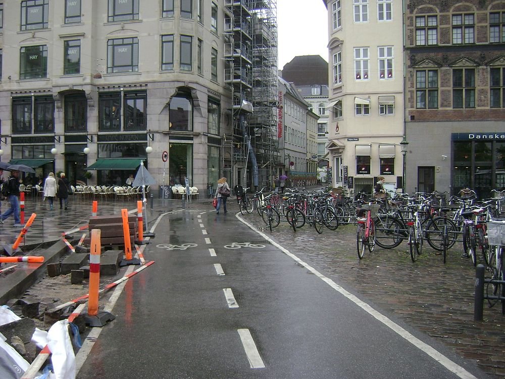 Regenachtige stedelijke straat met fietspad en geparkeerde fietsen Stedelijk straatbeeld met natte bestrating, fietspad, talrijke geparkeerde fietsen en voetgangers bij cafés en gebouwen in regenachtig weer