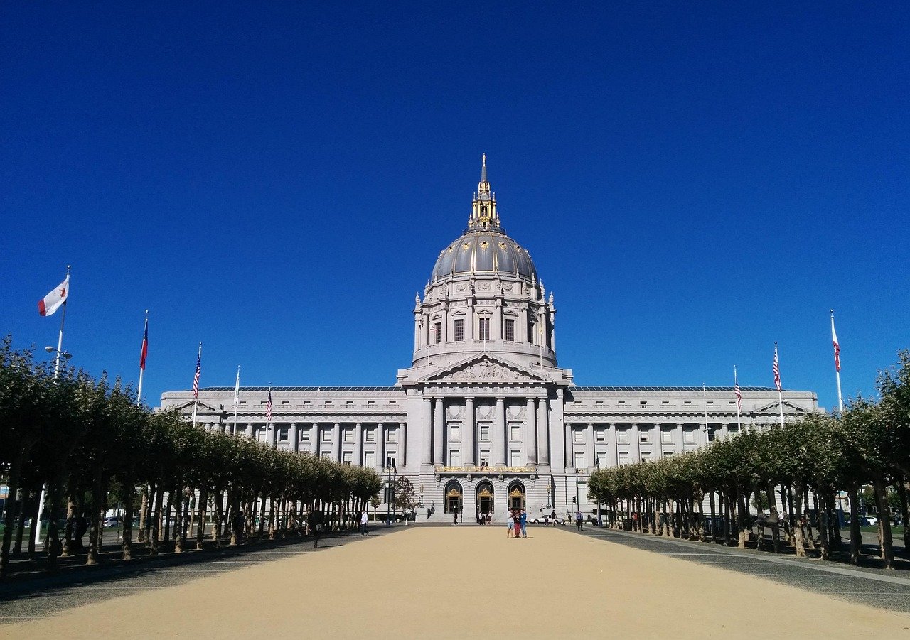 Stadhuis van San Francisco met grote koepel en vlaggen onder een heldere blauwe lucht, omgeven door bomen en een open plein