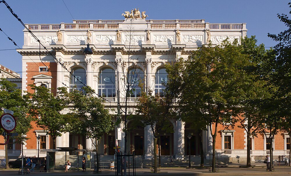 Historisch klassiek gebouw met sculpturen en bomen Historisch klassiek gebouw met sierlijke sculpturen en grote gebogen ramen achter groene bomen op een zonnige dag