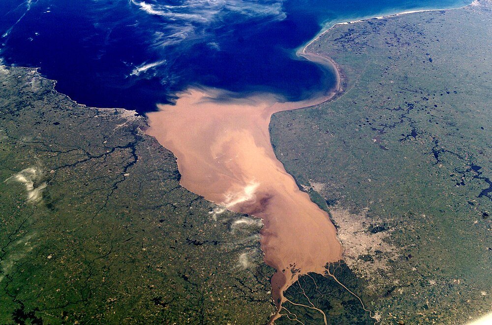 Luchtfoto van rivierdelta die de zee ontmoet Luchtfoto van een grote rivierdelta met modderig water dat in een diepblauwe zee stroomt, omgeven door groene en bruine landgebieden.
