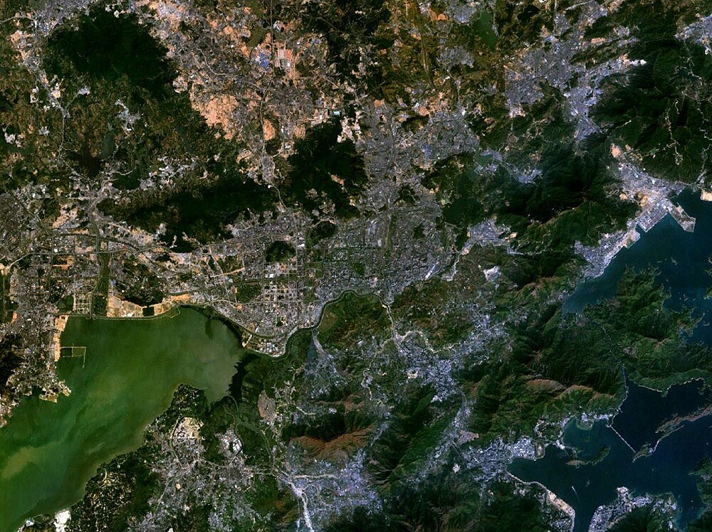Satellietbeeld van kuststad en omliggend landschap Satellietbeeld van een kuststad met stedelijke gebieden, groene bergen en waterlichamen, waaronder een baai en rivier