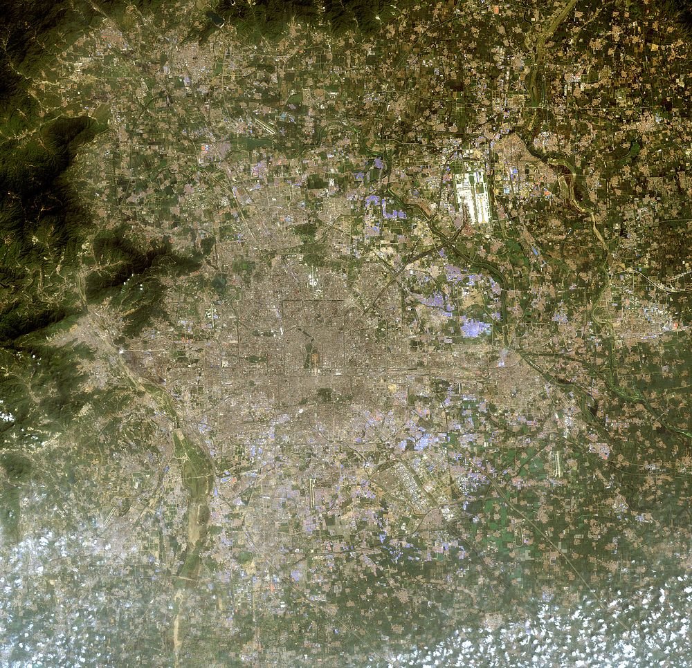 Satellietbeeld van stedelijke stad en omliggend landschap Satellietbeeld van een grote stedelijke stad omgeven door landbouwvelden en bergachtig terrein met verspreide wolken