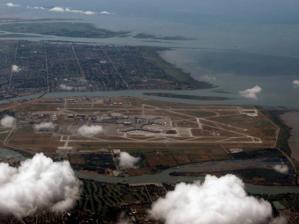 Luchtfoto van eilandluchthaven met start- en landingsbanen en terminals Luchtfoto van een grote luchthaven op een eiland omgeven door water en stedelijke gebieden met start- en landingsbanen en terminals zichtbaar