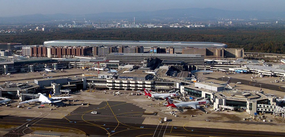 Luchtfoto van een drukke internationale luchthaven met meerdere vliegtuigen geparkeerd bij gates en terminalgebouwen zichtbaar