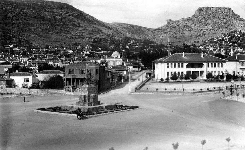 Historisch stadsplein met standbeeld en bergen Historische zwart-wit foto van een stadsplein met een standbeeld, traditionele gebouwen en bergachtige achtergrond