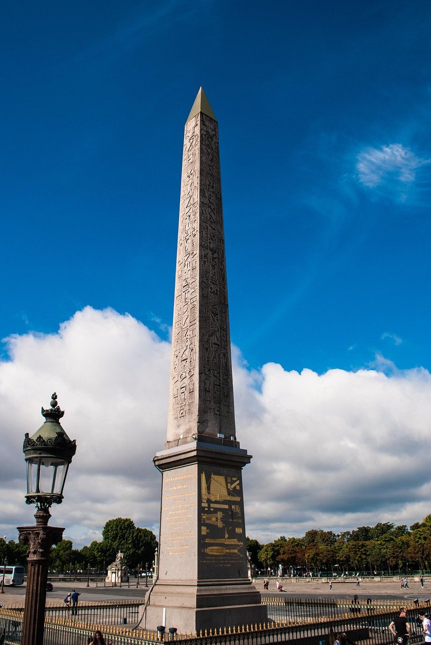 Hoge oude Egyptische obelisk met hiërogliefen op een openbaar plein onder blauwe lucht en witte wolken