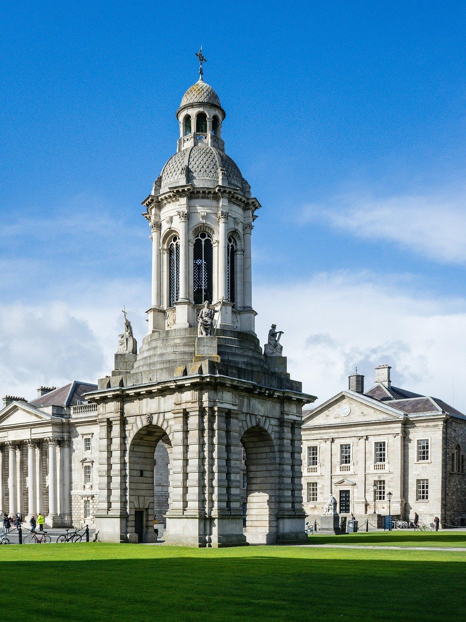 Historische stenen klokkentoren met beelden en bogen op de campus van Trinity College Dublin onder blauwe lucht