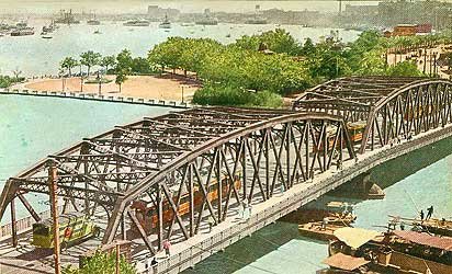 Historische stalen spoorbrug over rivier met treinen Historische stalen vakwerkspoorbrug over rivier met treinen en boten, omgeven door groen en stadsgezicht op de achtergrond