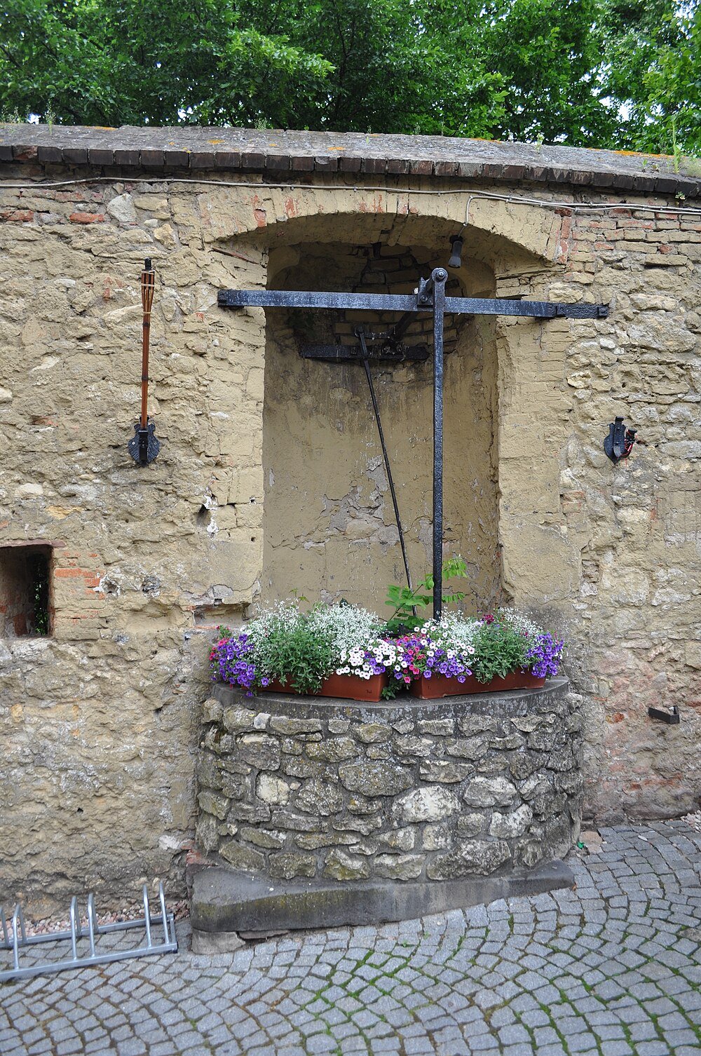 Oude stenen put met kleurrijke bloemen in potten bovenop, tegen een rustieke bakstenen en pleistermuur met groen erboven