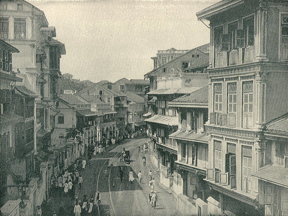 Historische zwart-wit foto van een drukke straat met koloniale gebouwen en mensen die lopen en paardrijden