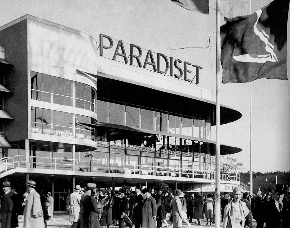 Historisch Paradiset-gebouw met menigte en vlaggen Zwart-wit foto van een menigte buiten het Paradiset-gebouw met grote glazen ramen en vlaggen op de voorgrond