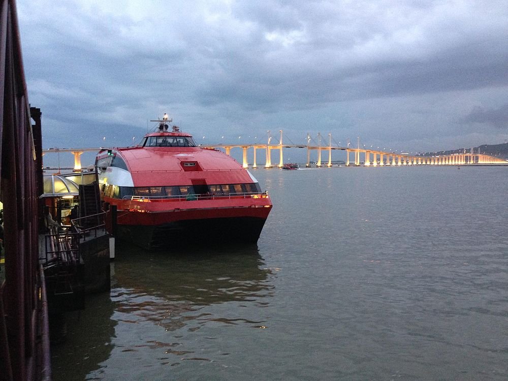 Rode en zwarte veerboot aangemeerd bij een pier met een lange verlichte brug op de achtergrond tijdens de schemering
