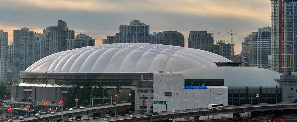 Wit koepelstadion met stadsgezicht en snelweg Groot wit koepelstadion met stadsgezicht en snelwegverkeer op de voorgrond tijdens zonsondergang