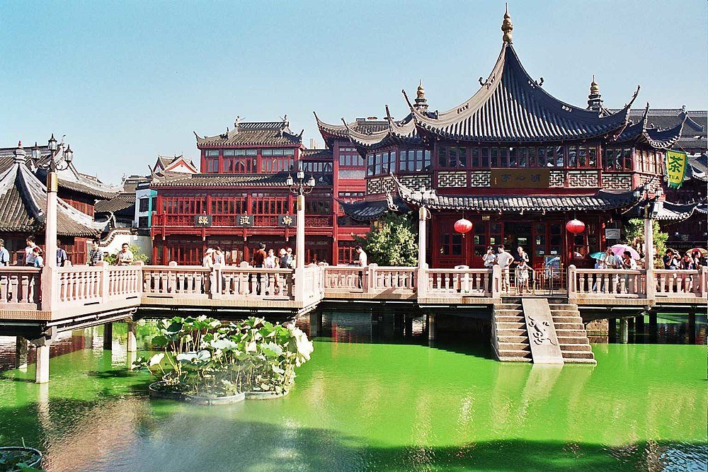 Traditionele Chinese gebouwen en stenen brug over vijver Traditionele Chinese architectuur met rode houten gebouwen en een stenen brug over een groene vijver met lotusplanten