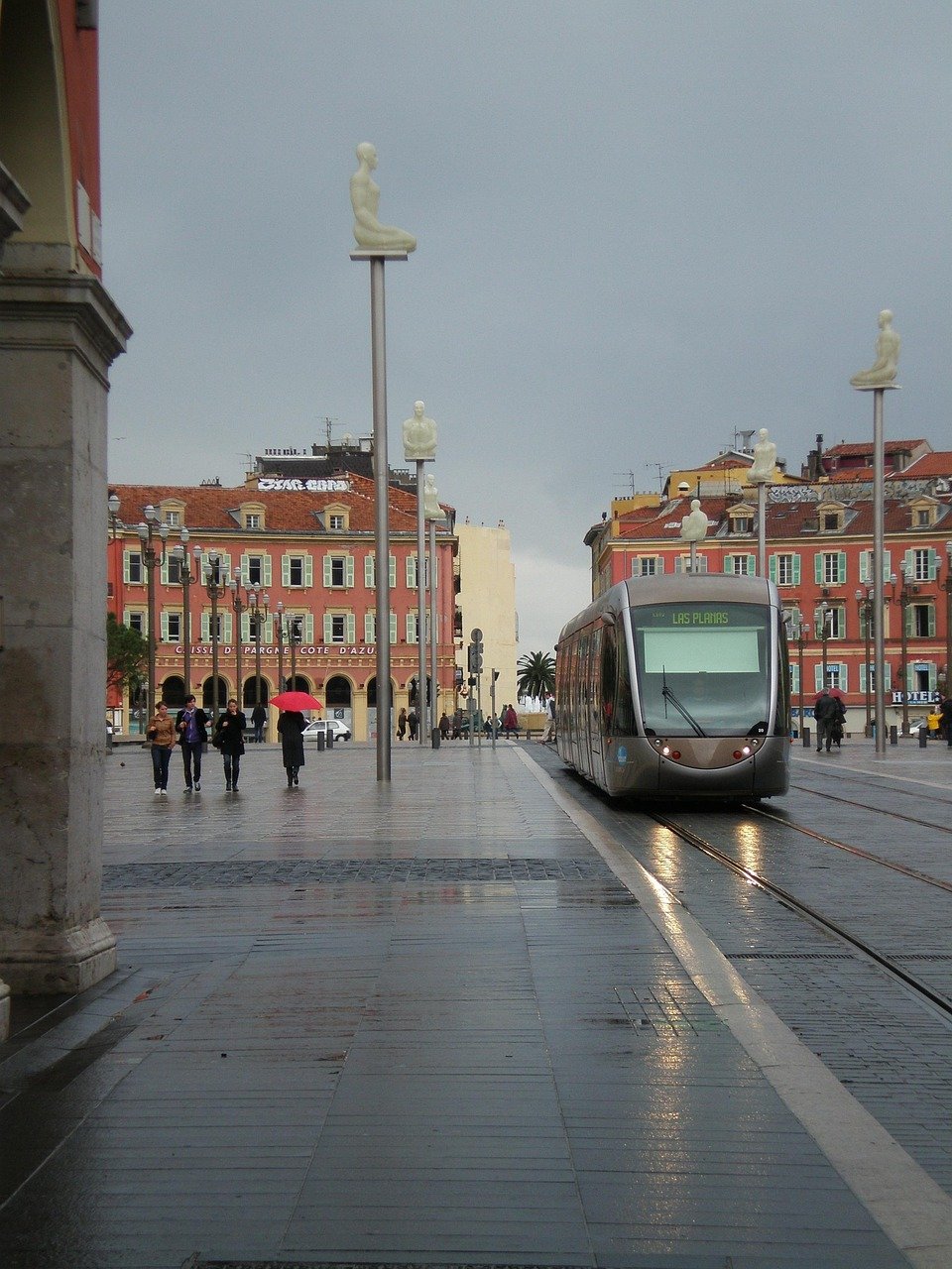 Moderne tram op natte straat met unieke zittende menselijke sculpturen op palen in een Europees stadsplein