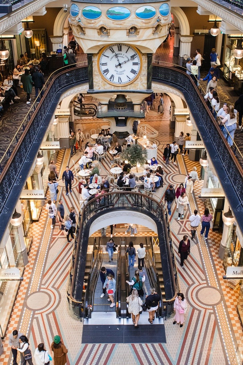 Interieur van een druk winkelcentrum met een grote klok, roltrappen en mensen die wandelen en dineren