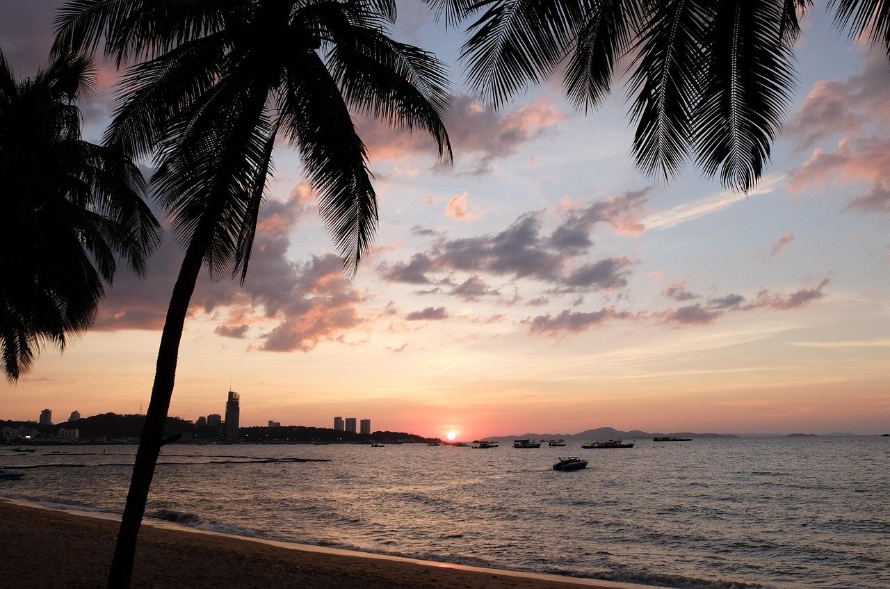 Zonsondergang boven een tropisch strand met palmbomen, kalme zee, boten en verre stadsgezicht onder kleurrijke lucht