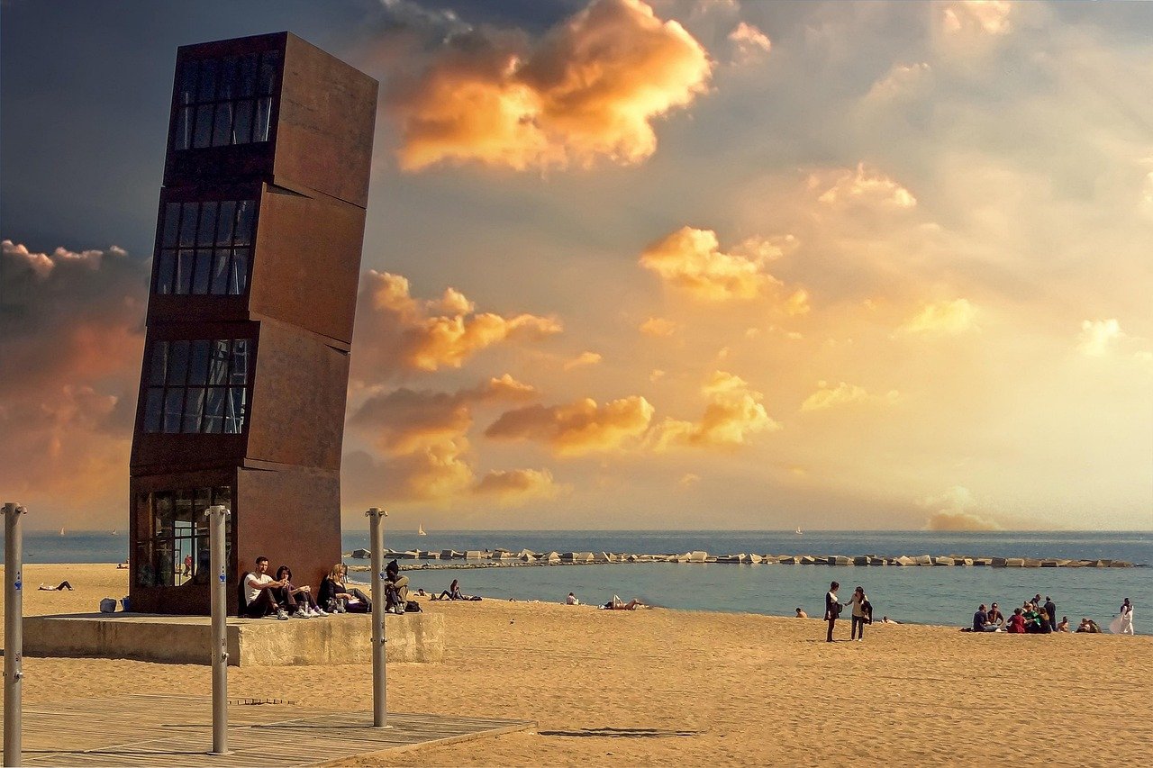 Moderne scheve toren sculptuur op een zandstrand met mensen die ontspannen en een levendige zonsondergang lucht boven de zee